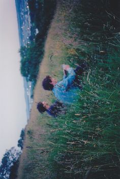 two people laying on the ground in grass next to a body of water and trees