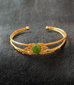 Vintage gold plated open bangle cuff bracelet with BC nephrite jade cabochon. Nephrite cabochon is 8 mm by 10 mm. The cabochon is mounted on a gold leaf and ribbon textural design. Bracelet arms are 3 mm thick. The balls at the inside ends of the cuff bracelet are 5 mm thick.  The bracelet is 60 mm thick. Nephrite jade is recognizable by its dark olive green, lightly translucent, colour. This jade was mined in British Columbia in the mid-1960s. Gold Jade Bangle Bracelet, Gold Cabochon Bangle Jewelry, Vintage Gold Bangle With Gemstone, Gold Jade Bangle, Handmade Elegant Jade Bangle, Formal Gold Jade Bracelets, Luxury Gold Jade Bracelets, Design Bracelet, Jade Bangle