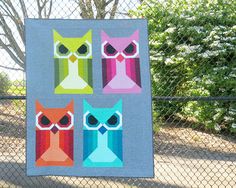 an owl themed quilt hanging on a chain link fence with trees in the back ground