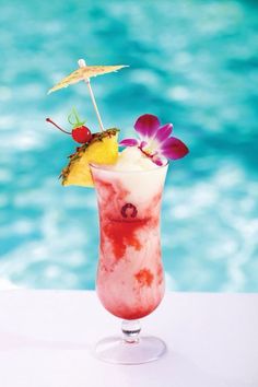 a drink with an umbrella and flowers in it sitting on a table next to a pool