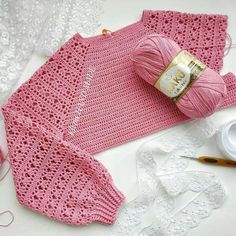 pink crocheted sweater next to yarn and scissors on white tablecloth with lace