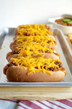 two hot dogs covered in cheese sitting on top of a metal baking pan next to a bowl of chili