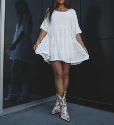 a woman standing in front of a black wall wearing white dress and cowboy booties