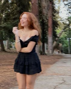 a woman in a short black dress is riding a skateboard on the sidewalk with trees behind her