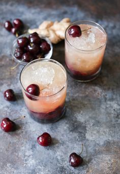 two glasses filled with ice and cherries on top of a stone table next to some fruit