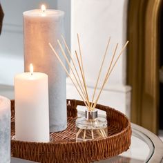 three candles are sitting on a wicker tray next to a glass candle holder with reeds in it