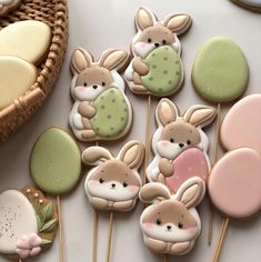 several decorated cookies are sitting on sticks next to a basket and cupcakes in the shape of bunnies