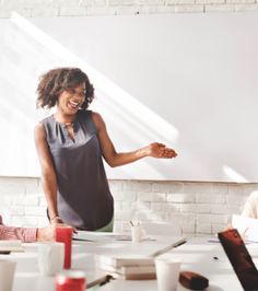 two women sitting at a table talking to each other in front of a whiteboard