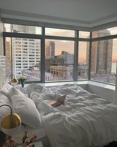 an unmade bed with white sheets and pillows in front of large windows overlooking the city