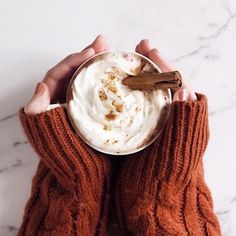 two hands holding a cup of hot chocolate with whipped cream and cinnamon sticks in it