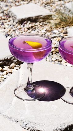 two glasses filled with purple liquid sitting on top of a stone slab next to rocks