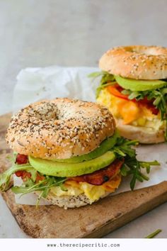 two bagel sandwiches with avocado and bacon on a cutting board next to each other