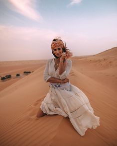 a woman sitting on top of a sand dune wearing a headdress and holding a cell phone to her ear