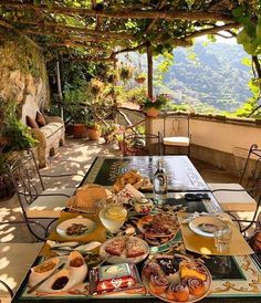 an outdoor table with food and drinks on it under a pergolated roof over looking the mountains