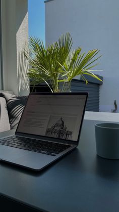 an open laptop computer sitting on top of a table next to a coffee cup and plant