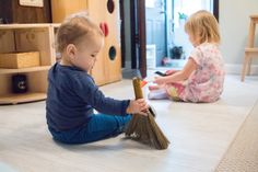 two children playing with toys on the floor in a living room, one holding a broom and the other sitting on the floor