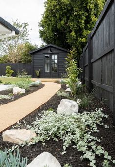 a garden with rocks and plants in front of a black fenced off area that is next to a house