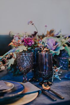 the table is set with two glasses and place settings, along with purple flowers in vases