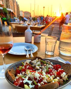 a plate of food on a table with wine glasses and people in the background at sunset