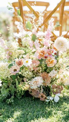 an arrangement of flowers is sitting on the grass in front of a wooden chair with greenery