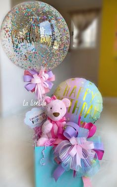 a teddy bear in a gift box with balloons and confetti on the table