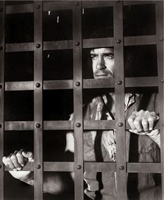 black and white photograph of a man in jail cell with his hand on the bars