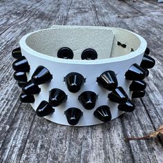 White leather studded punk bracelet with 3 rows of little cone studs. Studs measure 3/8" across and around 3/8" tall. Studs made with black enameled steel. Snaps made from matte black brass. Please note the the snaps have a nickel base layer. Cuff is 1.5" wide. Made with 7-8 oz (1/8", 3mm) white leather. Black Rock Style Jewelry With Studs, Rocker Style Leather Bracelet With Studs, Black Leather Jewelry With Studs, Adjustable Spiked Leather Bracelets, Edgy Black Bracelets With Studs, Edgy Adjustable Wristband With Studs, Black Leather Bracelet With Studs For Rock Style, Black Rocker Style Leather Bracelet With Studs, Rock Style Black Bracelets With Spikes