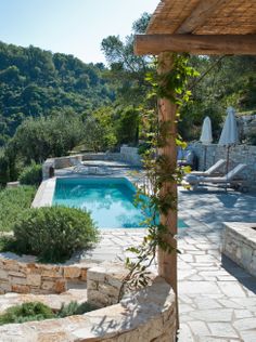 an outdoor swimming pool surrounded by stone walls and greenery next to a wooden pergolated gazebo