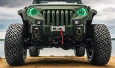 the front end of a green jeep parked on top of a sandy beach next to water