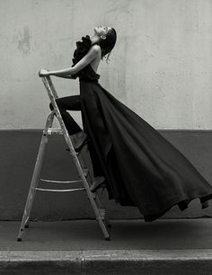 black and white photograph of woman in dress on ladder leaning up against wall with head tilted to the side