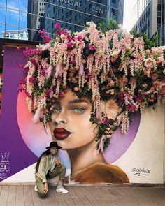 a woman sitting on the ground next to a wall with flowers growing over her head