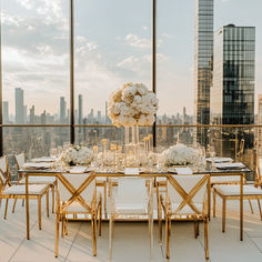 a dining table set with white flowers and gold chairs in front of large windows overlooking the city