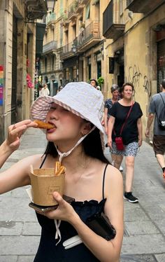 a woman in a hat eating food on the street with other people walking around her