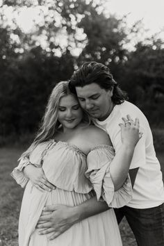 black and white photograph of a pregnant couple hugging each other in a field with trees in the background