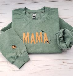 "Welcome to my store! This sweatshirt features an embroidered Spooky ooze font with the word \"mama\" in the middle of sweater, and a pumpkin on the right outer lower wrist. The thread color is orange but if you prefer a different color, kindly specify it in the personalization box. \"Stay cozy and stylish in this personalized embroidered sweatshirt! Made with high-quality material, this sweatshirt is designed to keep you warm and comfortable. The word 'mama' is beautifully embroidered on the front, making it a perfect gift for bridal showers or newlyweds.  This sweatshirt is perfect for a casual day out or a cozy night in. It comes in a range of sizes to fit anyone, and can be customized with your choice of trending hashtags to make it truly unique.  Make a statement with this mama sweats Green Fall Sweater With Embroidered Logo, Green Sweater With Embroidered Logo For Fall, Fall Green Sweater With Embroidered Logo, Green Embroidered Logo Sweater For Fall, Green Sweatshirt With Embroidered Logo For Fall, Long Sleeve T-shirt With Embroidered Logo For Fall, Custom Embroidered Long Sleeve Top For Halloween, Halloween Embroidered Long Sleeve Tops, Casual Halloween Top With Embroidered Logo