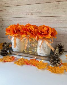 mason jars filled with orange flowers and pine cones are sitting on a tray surrounded by fall leaves