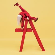 a woman in red jumpsuit sitting on top of a wooden ladder with her legs spread out