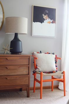 an orange chair next to a dresser with a lamp on it and pictures hanging above