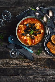 a bowl of stew with mussels and bread on a wooden table next to wine glasses