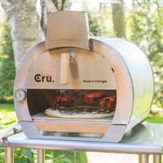 an outdoor pizza oven on a table in the sun with trees and grass behind it