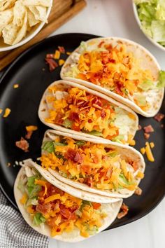 three tacos with shredded cheese and lettuce on a black plate next to bowls of chips