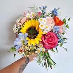 a person's hand holding a bouquet of colorful flowers and greenery on a white background