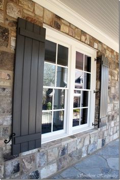 two windows with black shutters are on the side of a stone wall and brick building