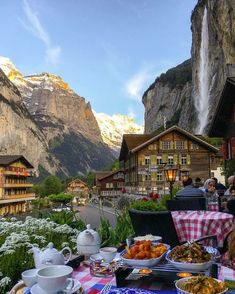 a table with food on it in front of some mountains and houses, near a waterfall