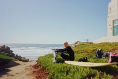 a woman sitting on the grass next to a surfboard near the ocean and a building