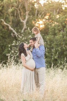 a man and woman holding a baby in their arms while standing in tall grass with trees in the background