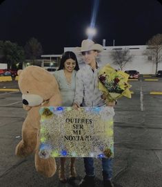 a man and woman standing next to a giant teddy bear holding flowers in front of a sign