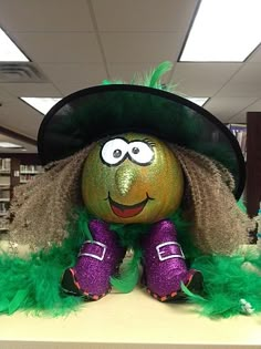 a green and purple stuffed animal sitting on top of a shelf in a library with bookshelves behind it