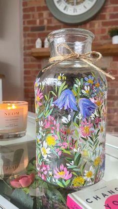 a glass jar with flowers painted on it sitting on a table next to a candle