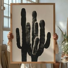 a woman holding up a black and white cactus art print in front of a window